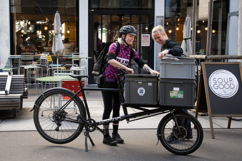 Kurierende und Mitarbeitende laden Kisten auf ein Lastenrad vor einem Café
